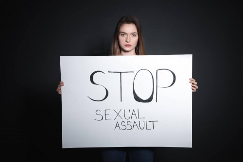 young-woman-holding-card-with-words-stop-sexual-assault-against-dark-background16eee76713a52525.jpg